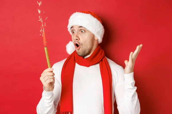 Conceito de Natal, férias de inverno e celebração. Close-up de homem bonito espantado em santa chapéu e cachecol, olhando para sparkler com emoção, de pé sobre fundo vermelho — Fotografia de Stock