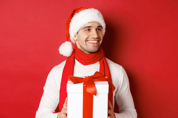 Концепция зимних праздников, Рождества и образа жизни. Крупный план привлекательного мужчины в шляпе Санты и шарфе, держащего новогодний подарок, смотрящего направо и улыбающегося, стоящего на красном фоне — стоковое фото