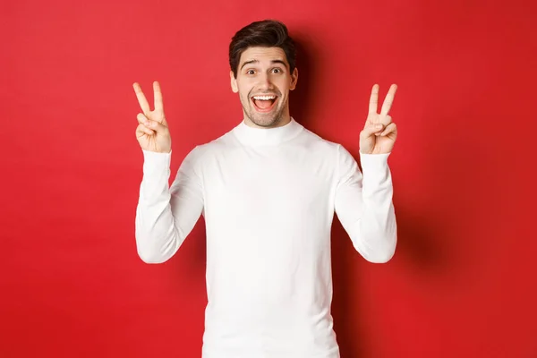 Concept of winter holidays, christmas and lifestyle. Handsome funny guy in white sweater, showing peace signs and smiling happy, standing against red background — Stock Photo, Image