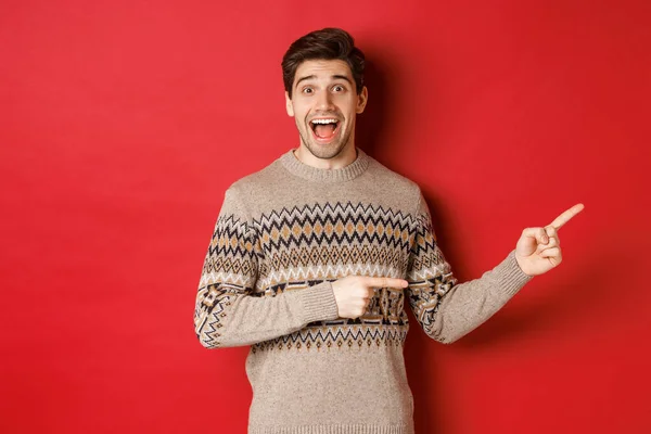 Concepto de celebración de Navidad, vacaciones de invierno y estilo de vida. Hombre guapo en suéter de Navidad señalando los dedos a la derecha, sonriendo sorprendido, mostrando la promoción de año nuevo contra el fondo rojo —  Fotos de Stock