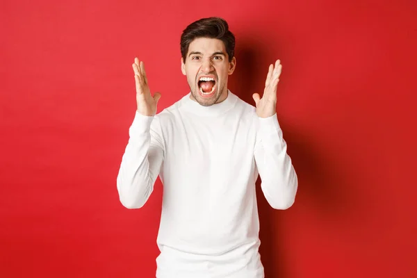 Obraz sfrustrowanego i wściekłego mężczyzny w białym swetrze, krzyczącego z wściekłości, wściekłego na kogoś, stojącego na czerwonym tle — Zdjęcie stockowe