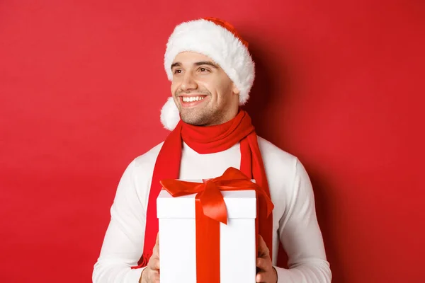 Concept de vacances d'hiver, Noël et style de vie. Gros plan de gars attrayant dans le chapeau et l'écharpe du Père Noël, souriant et regardant à gauche tout en tenant le nouvel an présent, debout sur fond rouge — Photo