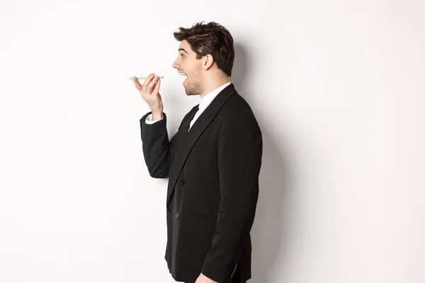 Profile shot of handsome businessman in black suit talking on speakerphone, smiling and looking happy, recording voice message, standing over white background — Stock Photo, Image