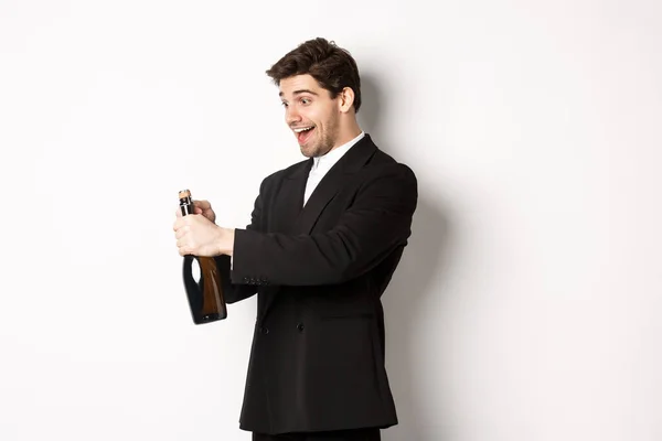 Retrato de homem atraente em terno preto, piscando para a câmera e abrindo garrafa de champanhe, celebrando o ano novo, de pé contra o fundo branco — Fotografia de Stock