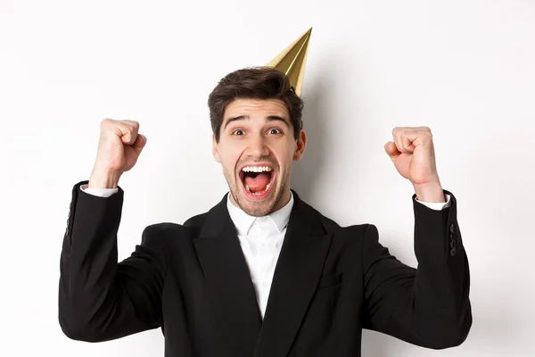 Close-up of happy good-looking man, wearing party hat and suit, raising hands up and rejoicing, celebrating new year, standing against white background — Stock Photo, Image