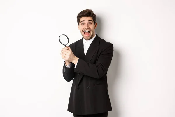 Handsome businessman in black suit, holding magnifying glass and smiling, found something, standing against white background — Stock Photo, Image