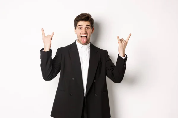 Portret van een vrolijke aantrekkelijke man die plezier heeft op een feestje, een zwart pak draagt, rock-n-roll teken en tong toont, opgewonden staat tegen een witte achtergrond — Stockfoto
