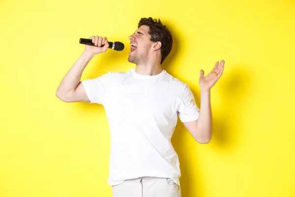 Jovem cantor segurando microfone, atingindo nota alta e karaoke cantando, de pé sobre fundo amarelo — Fotografia de Stock