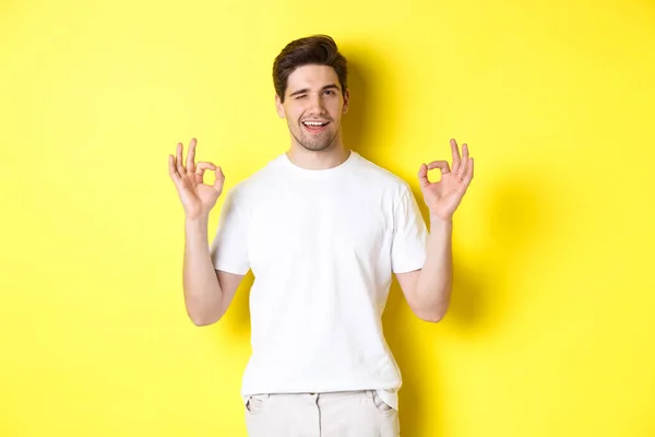 Zelfverzekerde knappe man knipoogt, toont goede tekenen in goedkeuring, als iets goeds, staande over gele achtergrond — Stockfoto