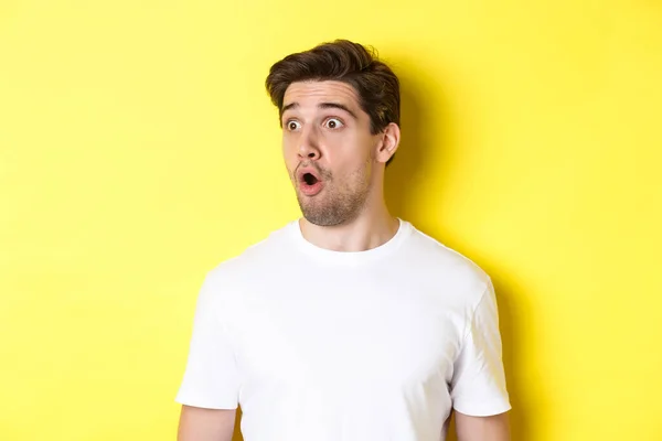 Close-up of impresated man looking left, gasping amazed, standing in white t-shirt against yellow background — Stok Foto