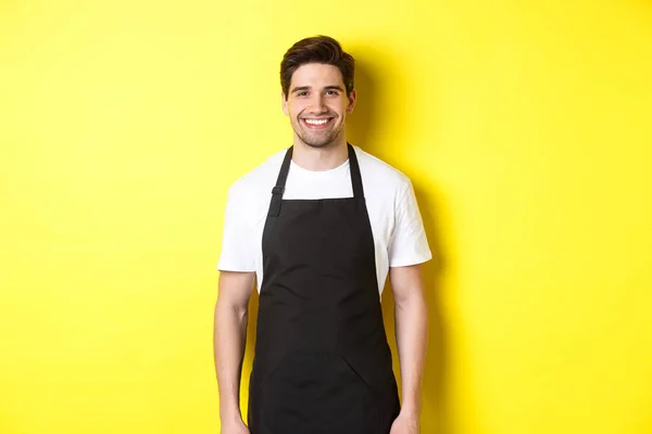 Jongeman ober in zwart schort, glimlachen, werken in de winkel of coffeeshop, staande tegen een gele achtergrond — Stockfoto