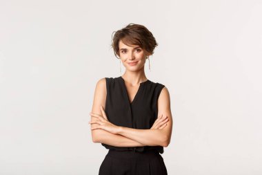 Zarif giysiler içinde duran ve gülümseyen genç ve zarif bir kadın. Hırslı iş kadını kameraya bakıyor, göğsünde çapraz kollar var.