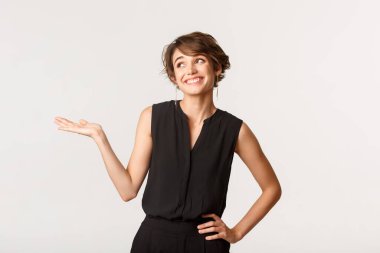 Göz kamaştırıcı genç bir kadın, yüzünü çevirmiş gülümsüyor, elini yana açıyor, ürününü beyaz arka planda tutuyor.