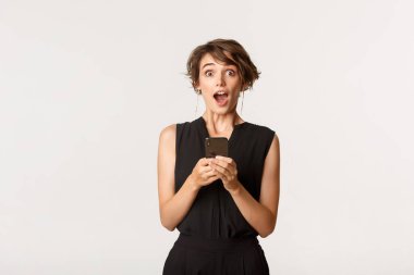 Şık elbiseli, akıllı telefon tutan, ağzı açık, mesajı okuduktan sonra şaşırmış, beyaz arka planda duran harika bir kadın.