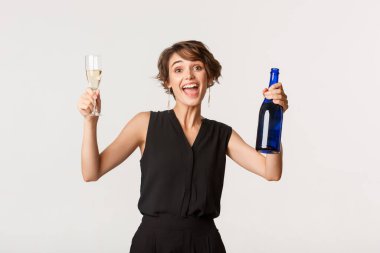 Mutlu kız eğleniyor, kutlama yapıyor, şampanya içiyor ve gülümsüyor, beyaz arka planda dikiliyor.