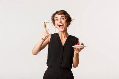 Neşeli doğum günü kızı elinde bir bardak şampanya ve yanan mumla beyaz arka planda duran bir parça pasta tutuyor.