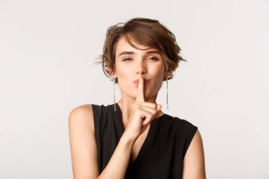 Şşş yapan, sırrı saklayan, dudaklarına parmak basan, beyaz arka planda duran zarif güzel bir kadının yakın çekimi.