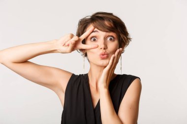 Aptal, güzel bir kadının somurtması ve barış jestini gözünün önünde göstermesi, beyaz arka planın karşısında durması.