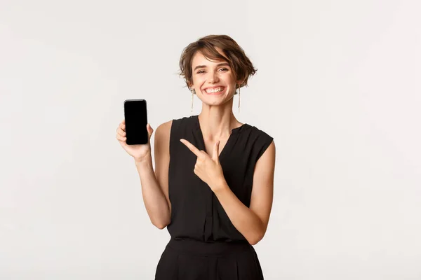 Šťastná stylová mladá žena s potěšeným úsměvem, ukazující prstem na displej mobilního telefonu, ukazující aplikaci, stojící bílé pozadí — Stock fotografie