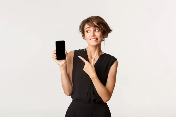 Oavgörande söt flicka peka finger på smartphone skärm och bita läpp tvekande, stående vit bakgrund — Stockfoto