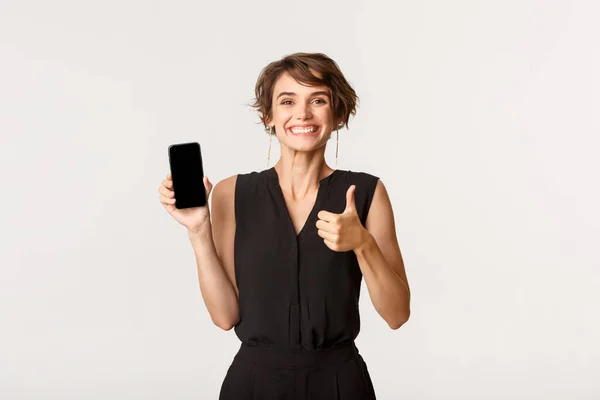 Nöjd vacker dam visar tummen upp i godkännande, visar smartphone skärm, stående vit bakgrund — Stockfoto