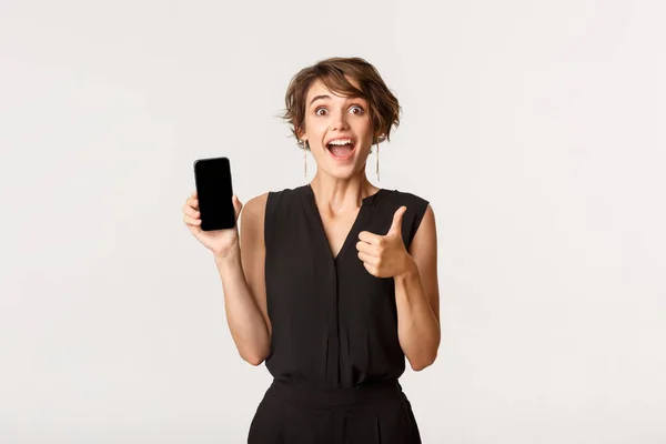 Úžasná mladá žena ukazující palce nahoru a mobilní telefon obrazovku, doporučit obchod nebo aplikaci, stojící bílé pozadí — Stock fotografie
