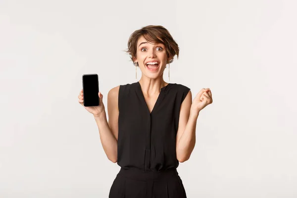 Spännande kvinna ser glad medan du visar smartphone skärm, leende roade, stående vit bakgrund — Stockfoto