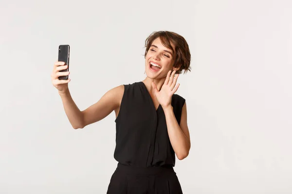 Веселая элегантная женщина здоровается во время видеозвонка, машет на камеру смартфона и улыбается, стоя на белом фоне — стоковое фото