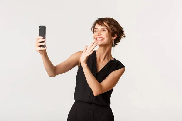 Счастливая шикарная женщина, имеющая видеозвонок, машущая рукой в камеру смартфона и улыбающаяся, стоя на белом фоне — стоковое фото