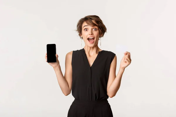 Úžasné roztomilé dívka otevřených úst fascinován, ukazuje kreditní karty a mobilní telefon obrazovku, stojící bílé pozadí — Stock fotografie