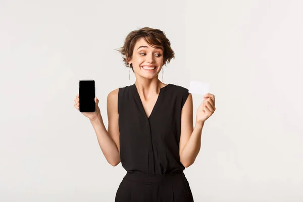 Spännande söt flicka ser glad på kreditkort, visar mobiltelefon skärm, stående vit bakgrund — Stockfoto
