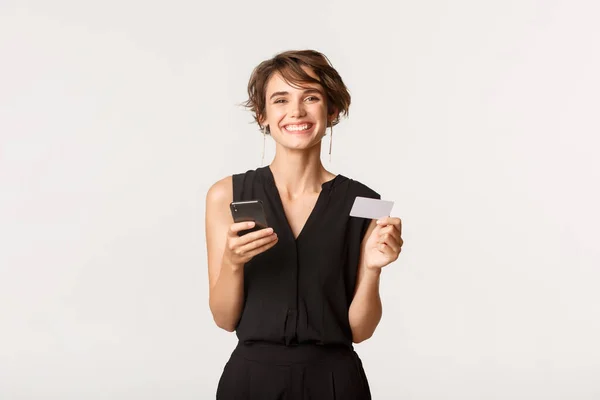 Alegre hermosa hembra con tarjeta de crédito y teléfono inteligente sonriendo a la cámara — Foto de Stock