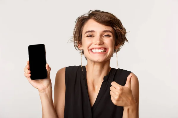 Bild av lycklig nöjd kvinna rekommendera något, visar tummen upp och smartphone skärm, står över vit bakgrund — Stockfoto