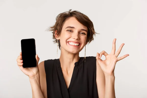 Obrázek veselé potěšené dívky ukazující dobré gesto, usmívající se a demonstrující obrazovku mobilního telefonu, stojící nad bílým pozadím — Stock fotografie