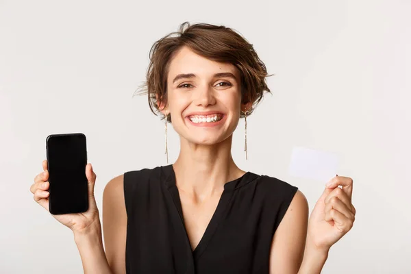 Detailní záběr spokojené usměvavé ženy ukazující kreditní kartu a displej mobilního telefonu, stojící nad bílým pozadím — Stock fotografie
