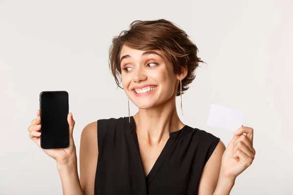 Detailní záběr krásné veselá mladá žena ukazující kreditní kartu, při pohledu na displej mobilního telefonu, stojící nad bílým pozadím — Stock fotografie