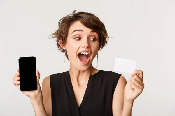 Close-up van mooie vrolijke jonge vrouw tonen mobiele telefoon scherm, kijken naar credit card enthousiast, staande over witte achtergrond — Stockfoto
