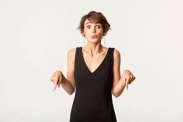 Immagine di eccitata donna bruna elegante che punta il dito verso il basso e guarda curiosa la fotocamera, in piedi su sfondo bianco — Foto Stock