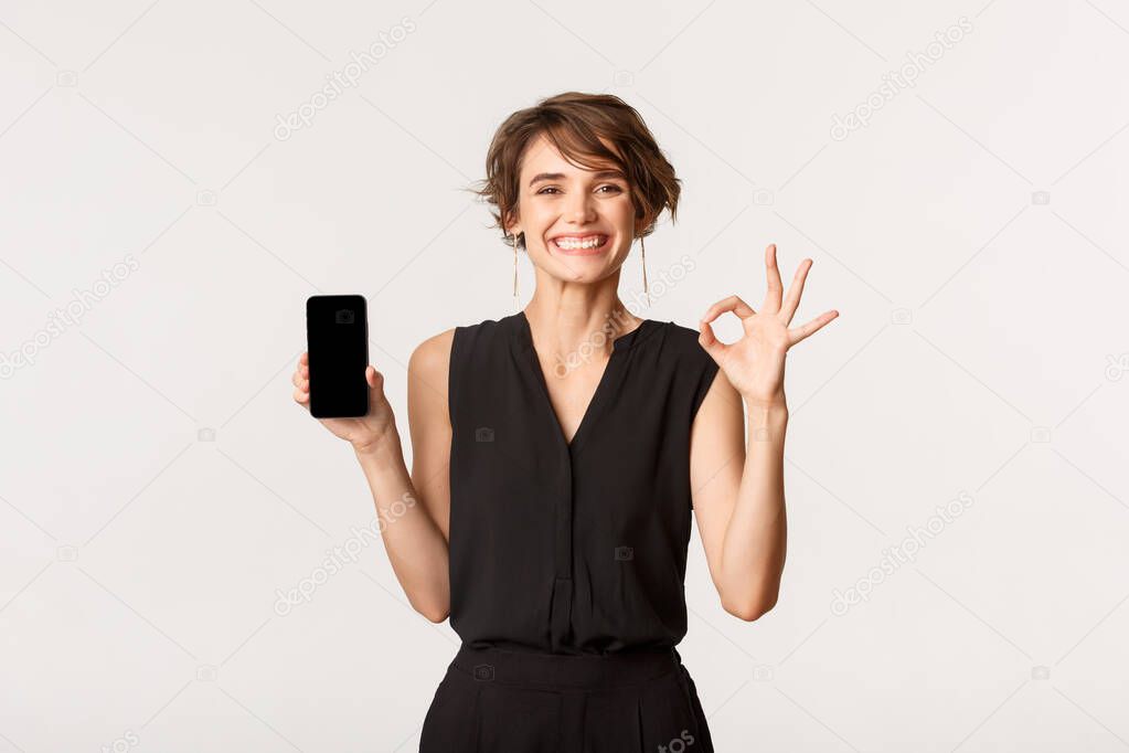 Satisfied young happy woman showing mobile phone screen and okay gesture, guarantee or recommend something, standing white background