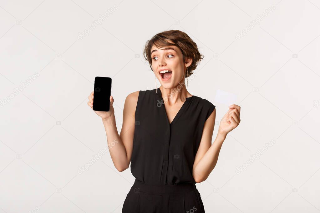 Amazed young woman showing credit card and looking at smartphone screen with intrigued smile, standing white background