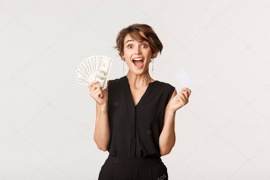 Excited lucky woman rejoicing, showing credit card and money, standing over white background