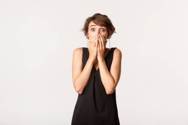 Şaşırmış genç bir kadın nefes nefese, elleriyle ağzını kapatıyor ve kameraya bakıyor beyaz arka planda dikiliyor.