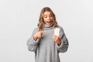 Gri kazaklı çekici beyaz kadın modelin portresi, şaşırmış ve cep telefonuna bakıyor, bir şey tavsiye ediyor, beyaz arka plan.