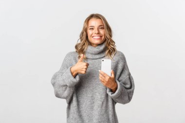 Gri kazaklı çekici sarışın kızın portresi tatmin olmuş görünüyor, baş parmağını kaldırıyor ve cep telefonu kullanıyor, bir şeyler öneriyor, beyaz arka planda duruyor.
