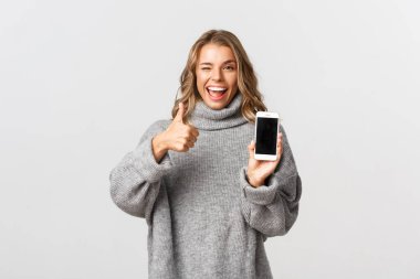 Gri kazaklı, çekici sarışın bir kız resmi. Tatmin olmuş görünüyor ve akıllı telefon ekranında güzel bir şey gösteriyor. Onaylanmış, beyaz bir arka plan.