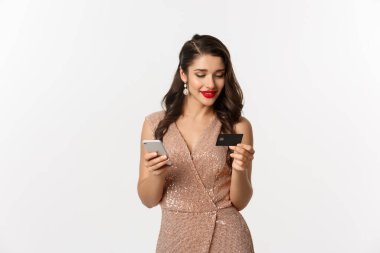 İnternetten alışveriş ve noel konsepti. Akıllı telefon ve kredi kartıyla internet alışverişi yapan zarif genç bir kadın, beyaz arka planda lüks elbiseler içinde duruyor.