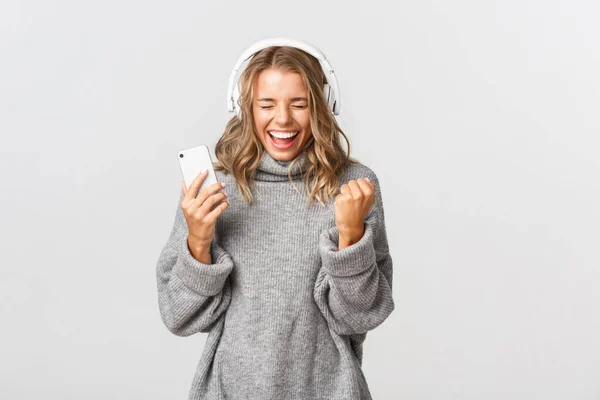 Söt vinnande flicka i grå tröja, lyssna musik i hörlurar, hålla smartphone och skriker av glädje, står över vit bakgrund — Stockfoto