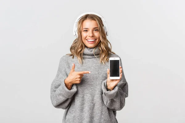 Studio bild av attraktiv blond flicka i grå tröja, lyssna musik i trådlösa hörlurar, visar smartphone skärm och leende, vit bakgrund — Stockfoto