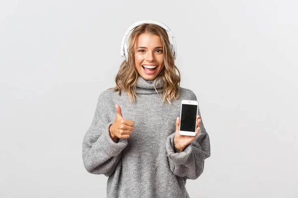 Imagem da menina loira satisfeita olhando feliz, mostrando polegares para cima, ouvir música em fones de ouvido e demonstrar tela do smartphone, fundo branco — Fotografia de Stock