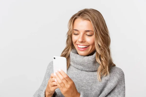 Närbild av attraktiv modern flicka med blond kort frisyr, bär grå tröja, med mobiltelefon och leende, står över vit bakgrund — Stockfoto